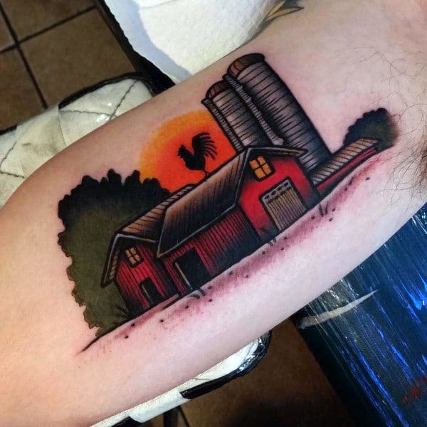 Vibrant color tattoo of a red barn with silos, a rooster on the roof, and a bold orange sunset in the background.