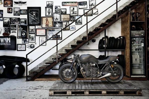 A vintage motorcycle sits on a wooden platform in a garage, nestled under a staircase with tire racks and framed photos