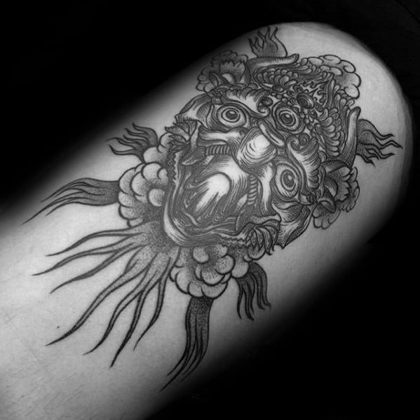 Garuda tattoo on the upper arm featuring a fierce face with ornate headgear, bulging eyes, flowing tendrils, and bold black shading.