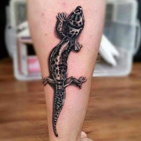 Realistic black and grey gecko tattoo on calf with detailed spots, shading, and a strong 3D effect that mimics a crawling lizard.