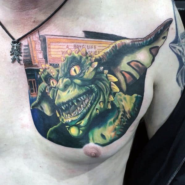 Realistic Gremlin chest tattoo with sharp teeth, glowing eyes, and a vibrant green hue, set against a nostalgic movie theater backdrop.