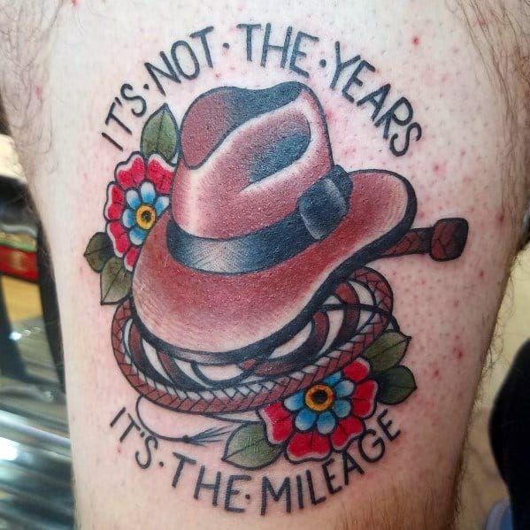 Colorful tattoo of Indiana Jones’ fedora and whip, surrounded by flowers, with the quote “It’s not the years, it’s the mileage.”