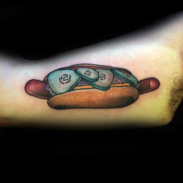 Tattoo of a hot dog featuring a sausage with pickles inside a bun, on the forearm.