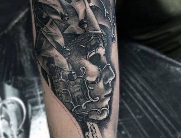 A black and gray tattoo of a skull with a jester hat, featuring intricate shading and detailing, placed on the forearm.
