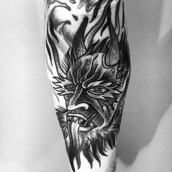 Black and grey Krampus tattoo with a fierce horned face, sharp teeth, intense eyes, and dynamic flame-like patterns surrounding the head.