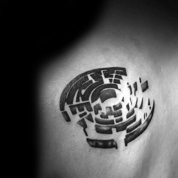 Abstract black ink labyrinth tattoo on a man's shoulder, featuring a fragmented circular maze with a 3D effect and scattered broken pathways.