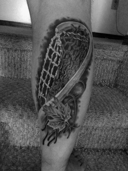 Realistic black and grey lacrosse stick tattoo with intricate netting, maple leaves, and bold shading, placed on the calf for a striking look.