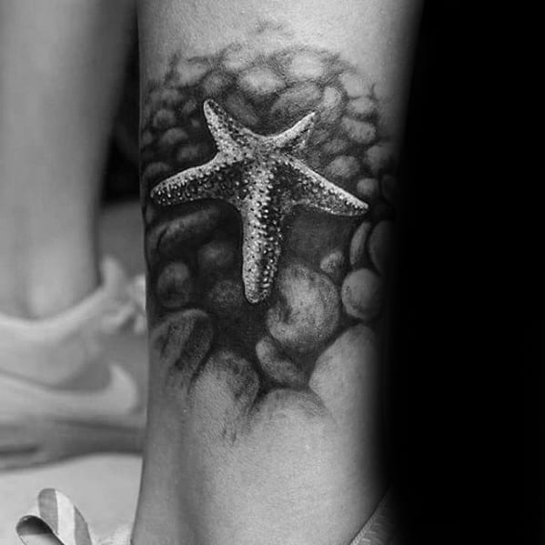 Realistic black and grey starfish tattoo resting on textured rocks, with strong shading to create a 3D ocean floor effect.