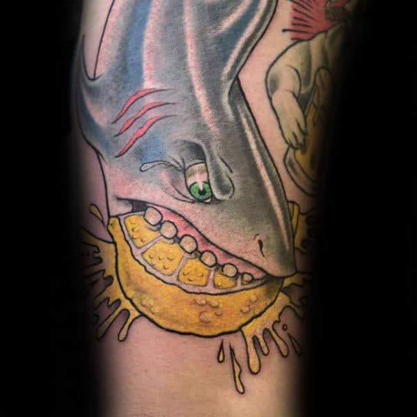 Bold and colorful tattoo of a shark biting into a juicy lemon, with exaggerated teeth and splashes of juice for a surreal, cartoonish effect.