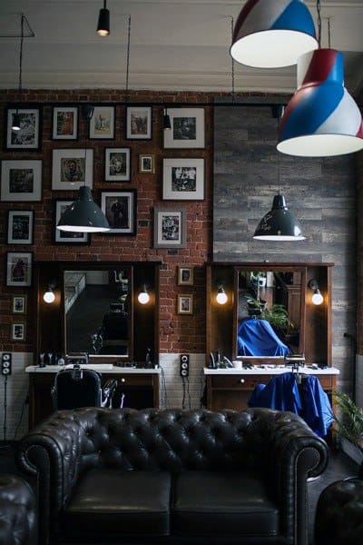 Industrial barbershop with leather sofa, brick walls, framed art, and pendant lighting.