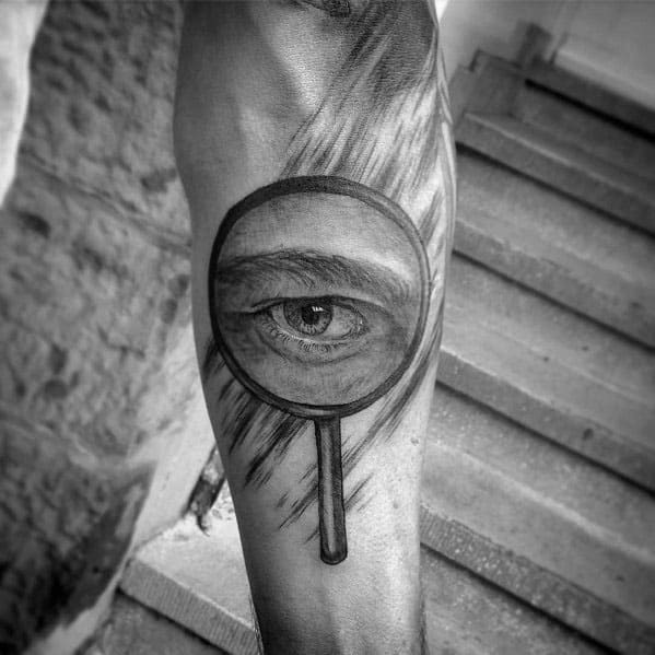 Tattoo of a magnifying glass focusing on a realistic eye, designed with detailed shading and depth for a striking and surreal visual effect.