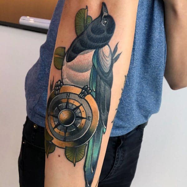 Colorful magpie tattoo on forearm, featuring a detailed bird perched on a compass with green leaves, symbolizing guidance and direction.