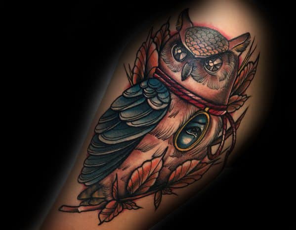Neo-traditional owl tattoo with rich shading, deep blue feathers, and an ornate eye pendant, surrounded by autumn leaves for a mystical touch.