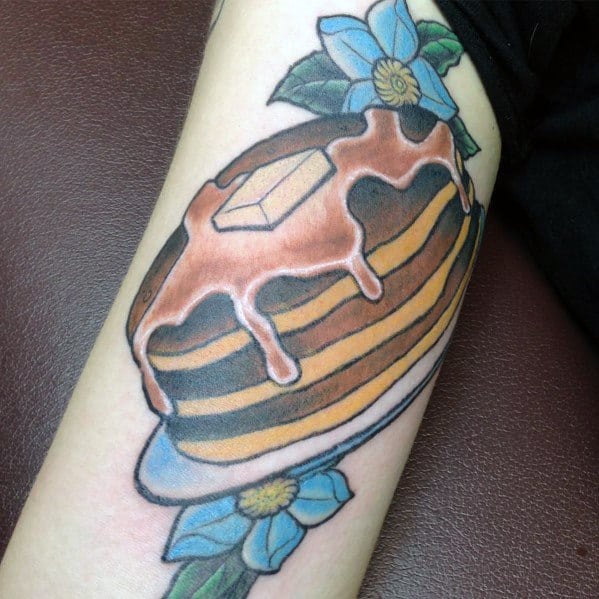 Realistic pancake tattoo with dripping syrup, butter, and blue flowers, featuring bold shading and vibrant colors for a decorative touch.