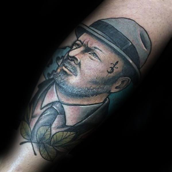 Colorful tattoo of a man in a fedora and suit, featuring a facial symbol and leaf accents on the forearm