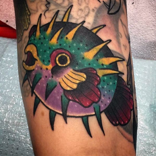 Colorful pufferfish tattoo with bold black outlines, vibrant green, yellow, and purple hues, and sharp spikes for a striking traditional look.