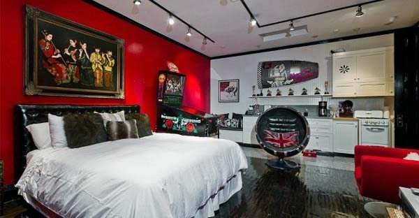 Stylish bedroom with a white bed, bold red and black decor, pinball machine, kitchenette, and retro chair
