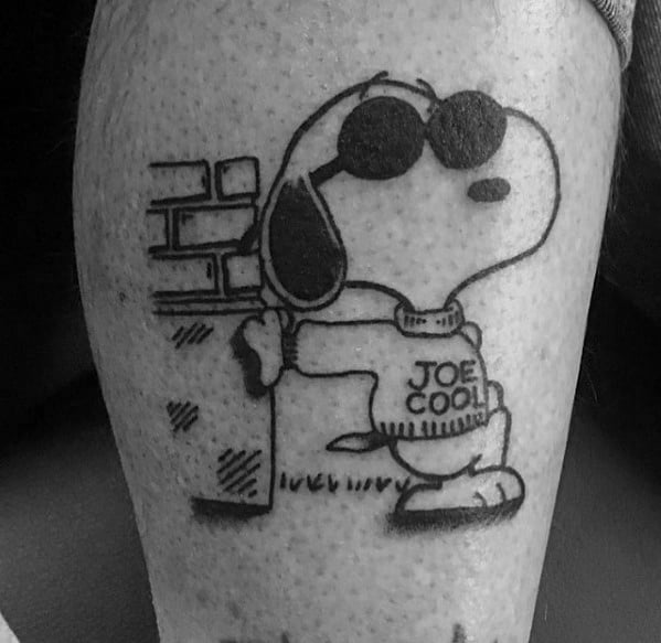 Black and white tattoo of Snoopy as "Joe Cool," wearing sunglasses and a sweater, leaning against a brick wall with a relaxed pose.