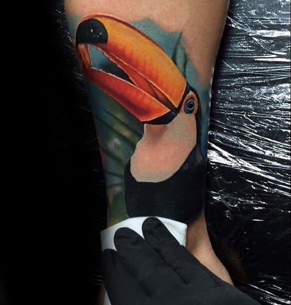 Hyper-realistic toucan tattoo with a vivid orange beak and lush green background, placed on the forearm for a striking nature effect.