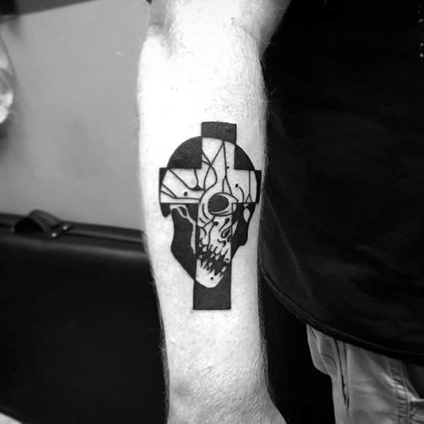 Black cross tattoo featuring a detailed ram skull in the center with sharp lines and bold shading, located on the forearm.
