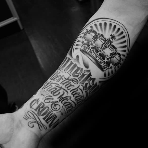 Forearm tattoo featuring a crowned emblem with radiant shading and bold script that reads "Uneasy lies the head that wears a crown."