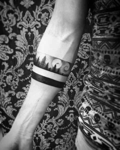 Minimalist black and grey forearm band tattoo featuring a forest silhouette with a moon and swirling clouds, paired with a bold solid black band.