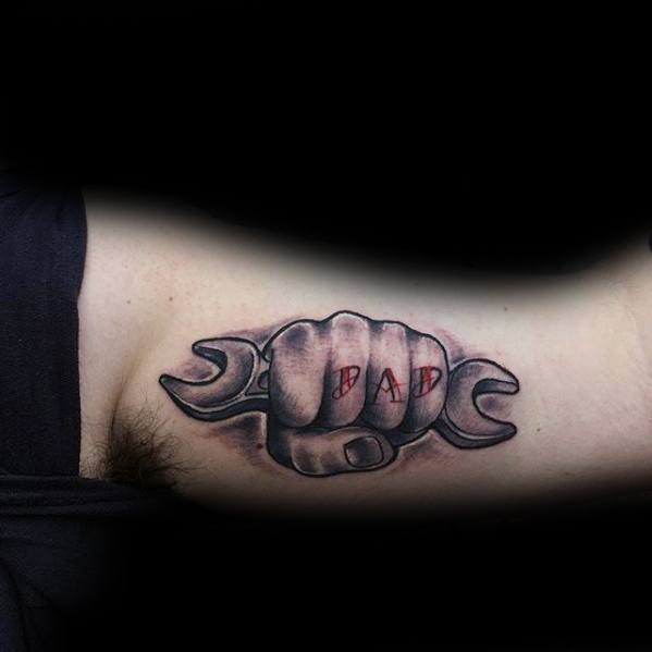 Tattoo of a fist gripping a wrench with the word DAD in red letters across the knuckles, shaded for a 3D look on the inner arm.
