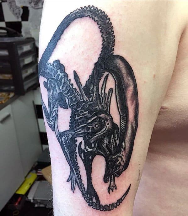 Black ink Xenomorph tattoo on upper arm showing the creature in a dynamic curled pose with a long, looping tail and sharp details.