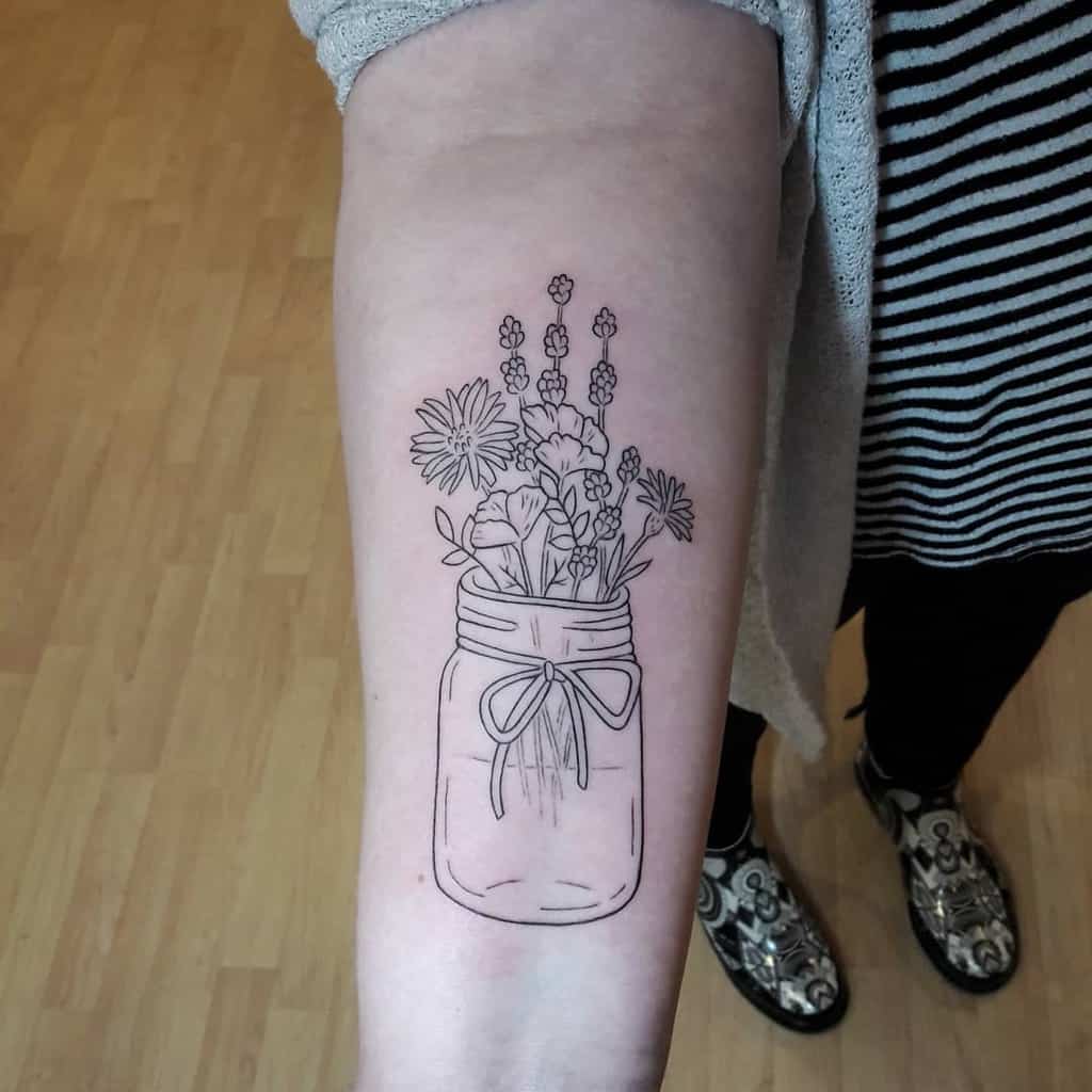 A tattoo on the upper arm depicting a jar filled with flowers, including lavender and daisies. The design is in black ink, featuring delicate outlines and a bow on the jar. The tattoo has a minimalistic and whimsical style.