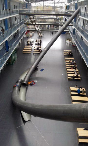 A sleek black slide winds through a spacious, modern building with people gathered at tables