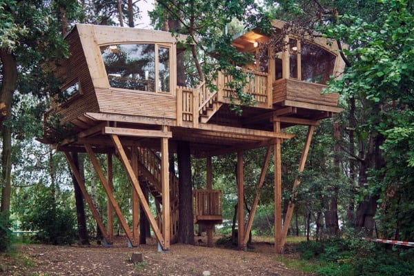 Modern wooden treehouse with large windows, elevated on stilts among green trees in a forest setting