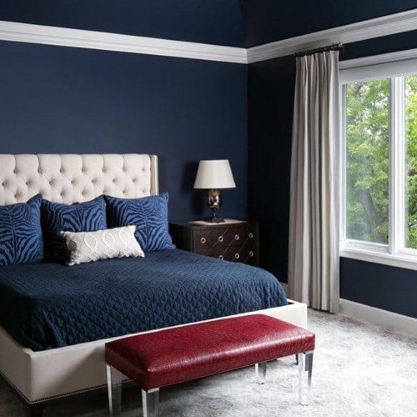 Elegant navy bedroom with tufted headboard, red bench, and large window with beige curtains.