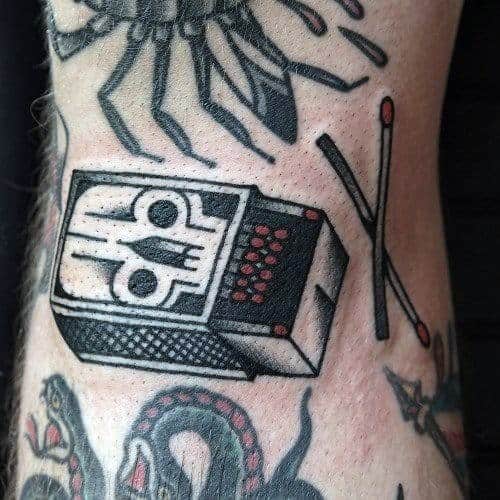 Traditional-style matchbox tattoo with a skull design and two red-tipped matches, symbolizing danger, rebellion, or fiery spirit.