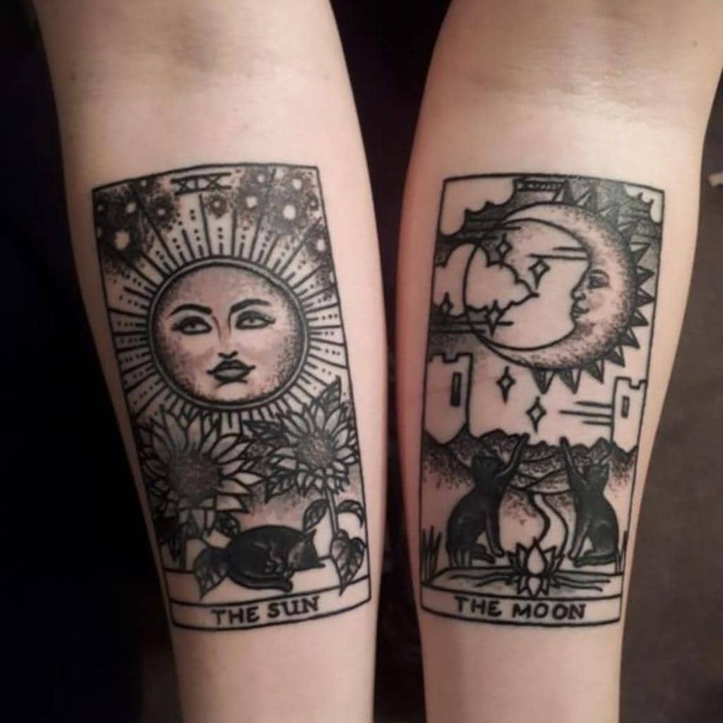 Matching sister tarot-style tattoos featuring 'The Sun' with a radiant face, sunflowers, and a sleeping cat, and 'The Moon' with a crescent moon, stars, and two cats around a fire