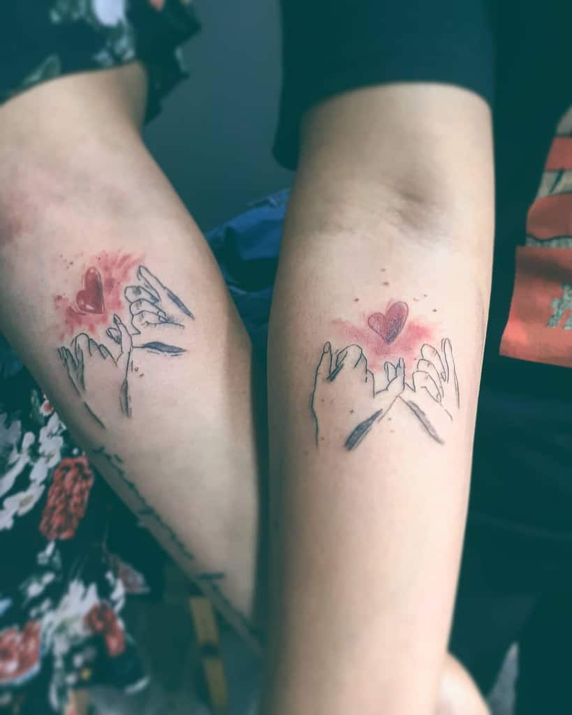 Two arms showing matching tattoos of hands making a pinky promise, with red hearts above each design