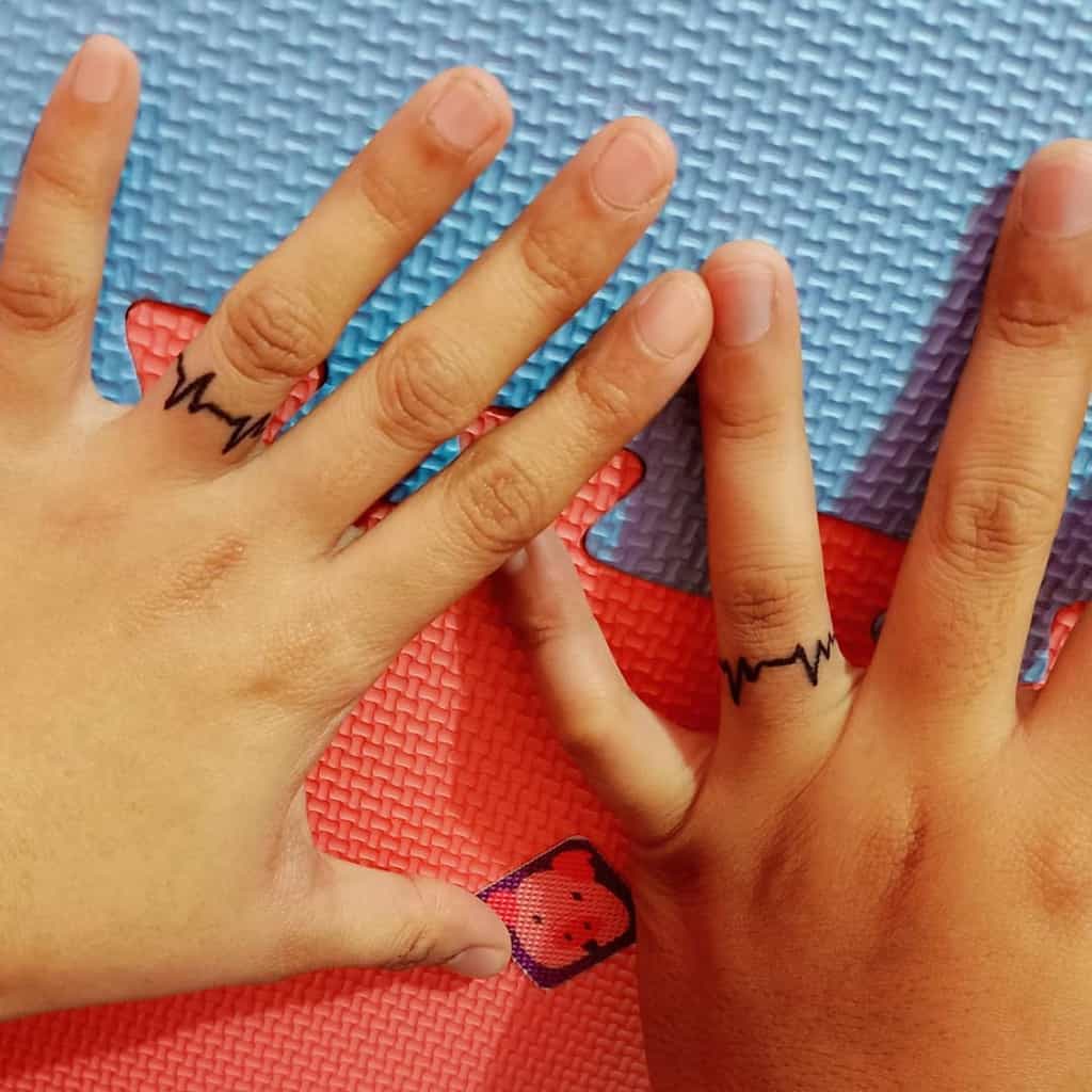 Matching heartbeat wedding ring tattoos on the fingers, symbolizing a strong and vital connection between the two individual