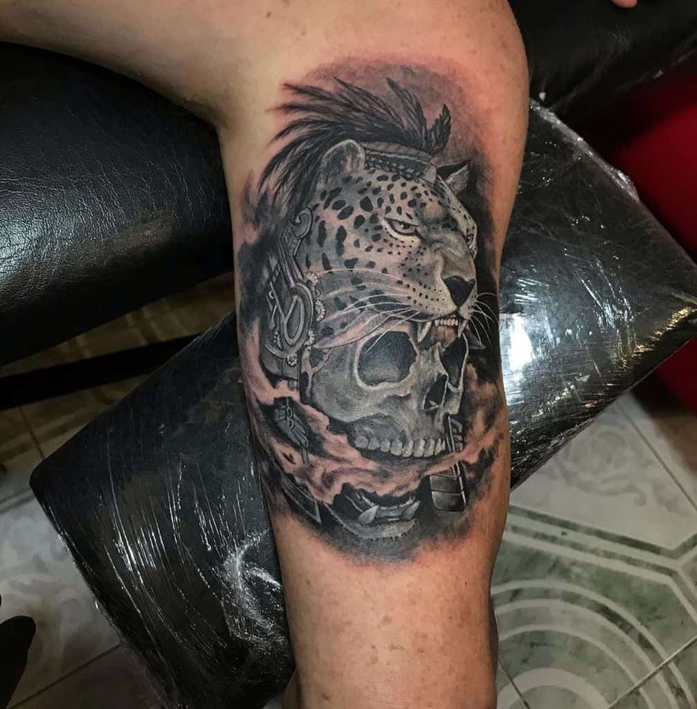 A powerful black and grey tattoo of a jaguar with a mohawk, seemingly about to devour a skull from which smoke is rising, with intricate shading and dramatic elements on the upper arm.