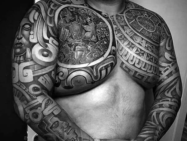 Black and grey Aztec-inspired tattoo covering the chest and arms, featuring intricate tribal patterns, deities, and ancient calendar designs.