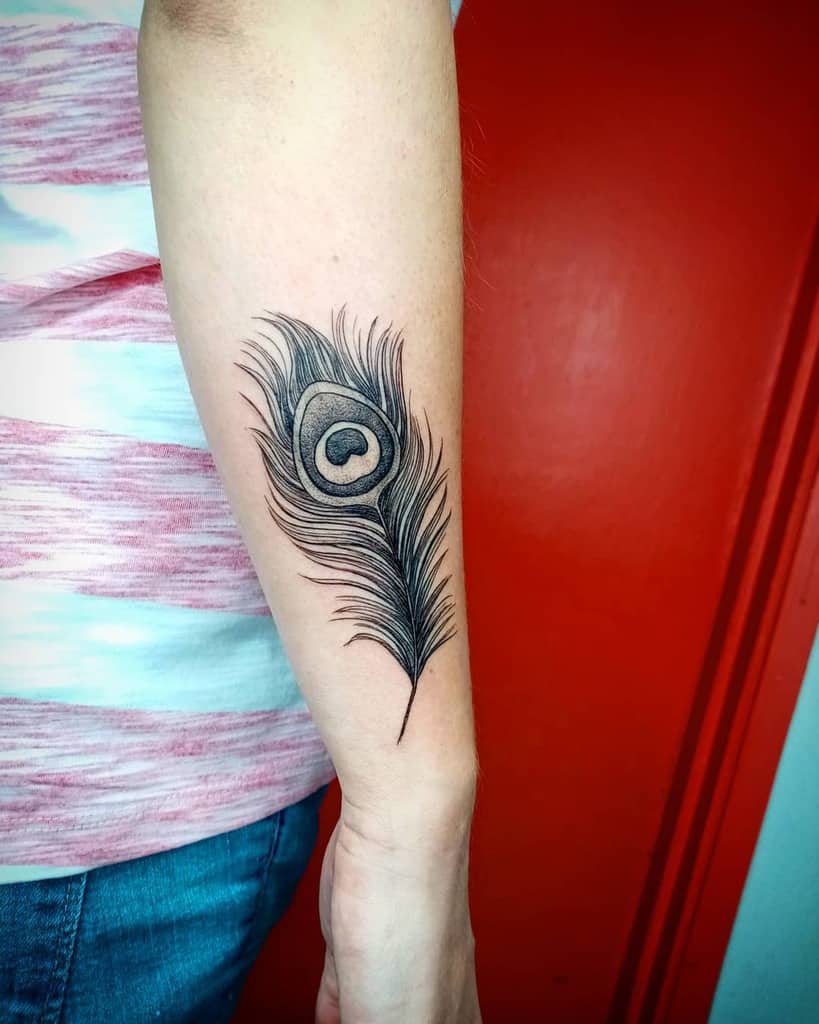 Black ink peacock feather tattoo on forearm, featuring fine lines and detailed shading with a bold eye pattern in the center.