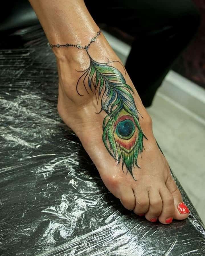 Colorful peacock feather tattoo on the foot, featuring rich green, blue, and gold tones with a beaded anklet design for added detail.