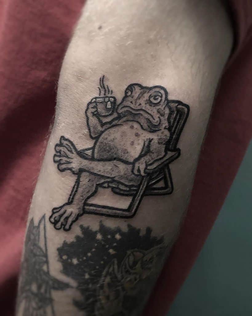 A black ink tattoo of a relaxed frog sitting in a chair, sipping a hot drink, drawn in a humorous and detailed style on the arm.