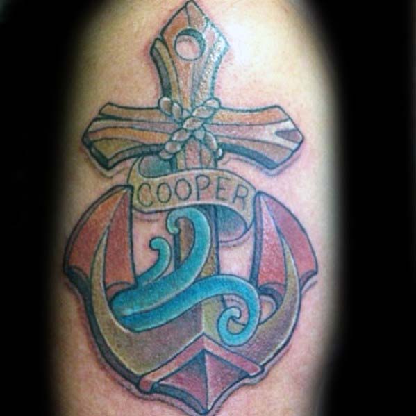 Tattoo of an anchor with a cross and ribbon saying "COOPER" on a person's skin