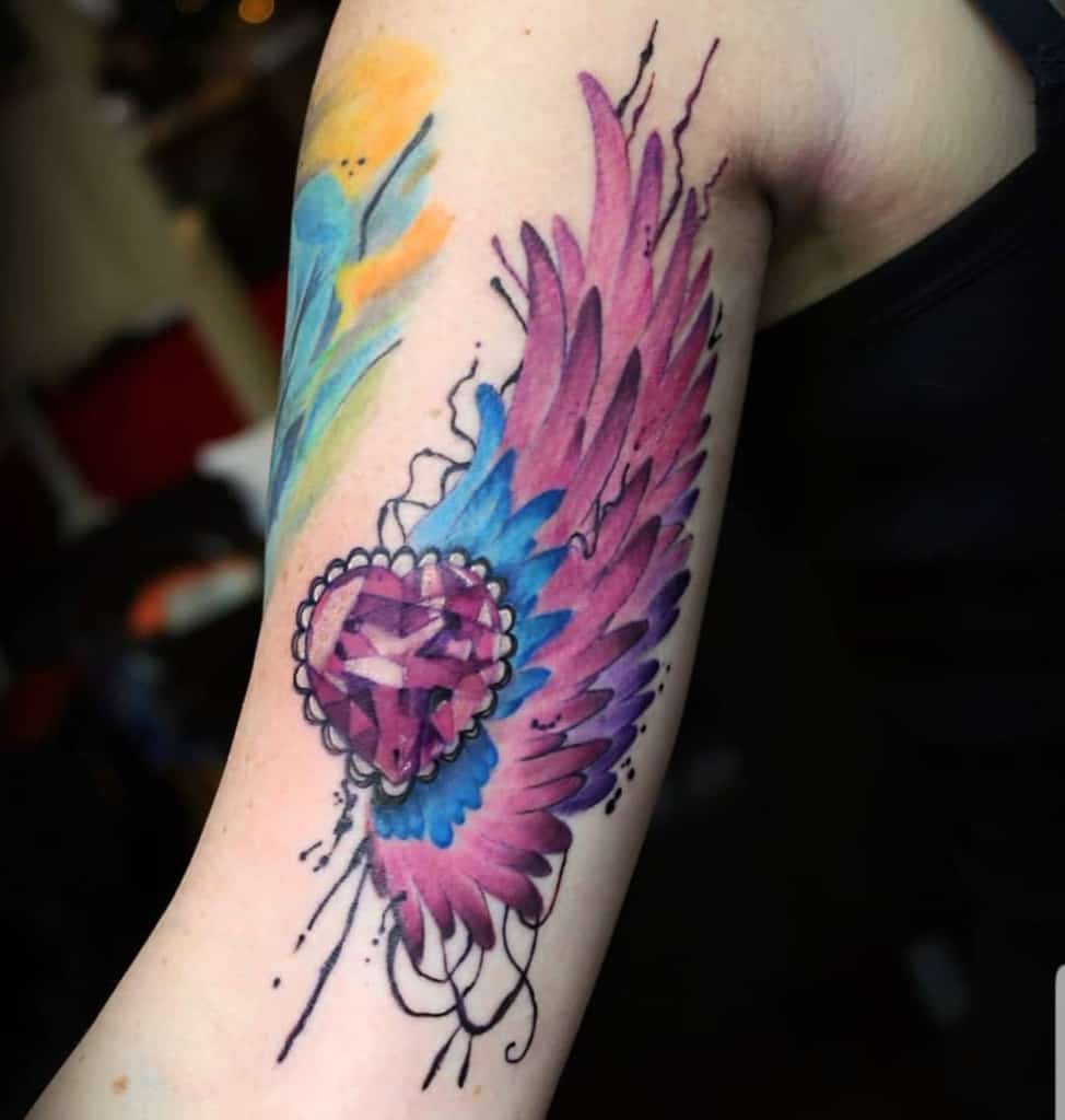 Vibrant watercolor-style tattoo of a pink gemstone with blue and pink angel wings, accented by abstract splashes of color