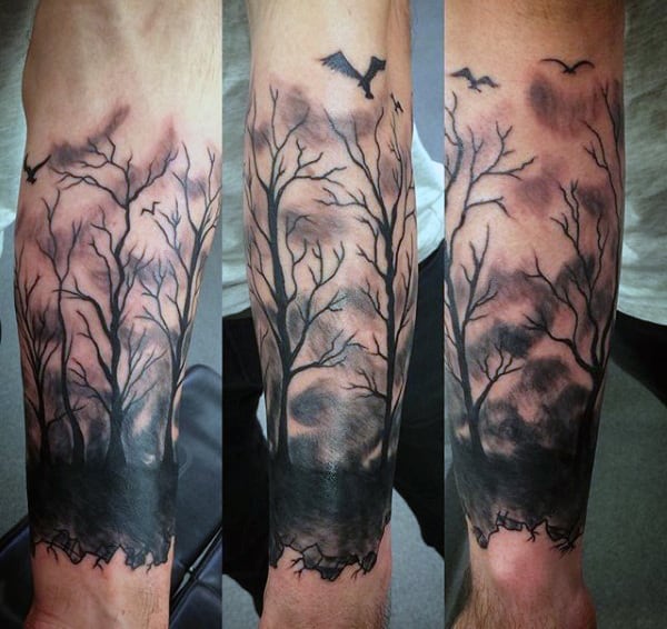 Forearm tattoo of a dark forest scene with bare trees and flying birds against a cloudy sky