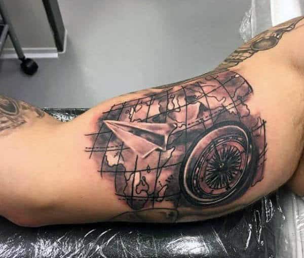 Arm tattoo of a paper plane, compass, and map in black and grey ink with a subtly integrated airplane motif