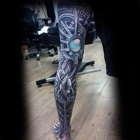 Leg tattoo with intricate biomechanical design, featuring metallic elements and a teal glowing orb, on a wooden floor background