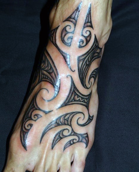 A tribal-style black tattoo covers the top of a foot with intricate swirling patterns and geometric shapes