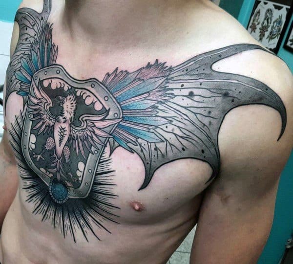 Chest tattoo of a winged shield with a phoenix-like creature, accented with blue and pink feathers, and bold black rays radiating below.