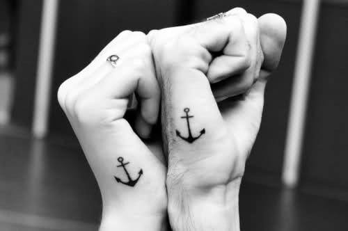 Matching side hand tattoos for men featuring small black anchors placed near the base of the thumbs.