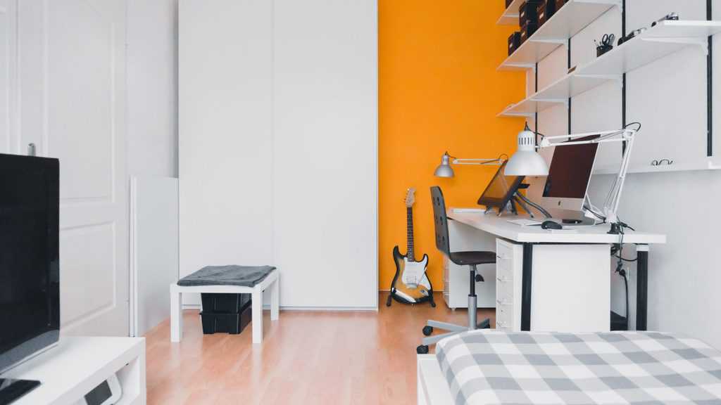 White minimalist bedroom with orange feature wall, work desk, and guitar decor.